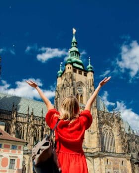 Woman in red dress looking up at a tall building with her arms up wide